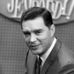 Art Fleming in a suit and tie hosting the original Jeopardy! show in 1964, standing behind a podium and holding cards.