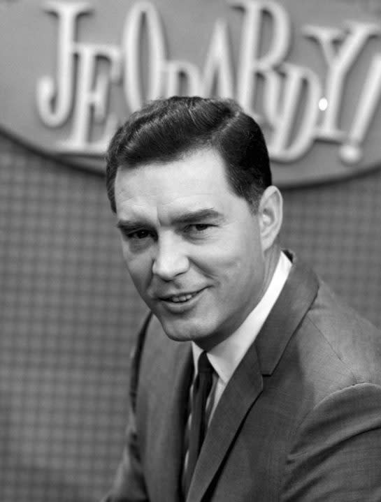 Art Fleming in a suit and tie hosting the original Jeopardy! show in 1964, standing behind a podium and holding cards.