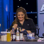 A chef using a spoon to remove something from a jar; the table has several bottles and cooking implements on it