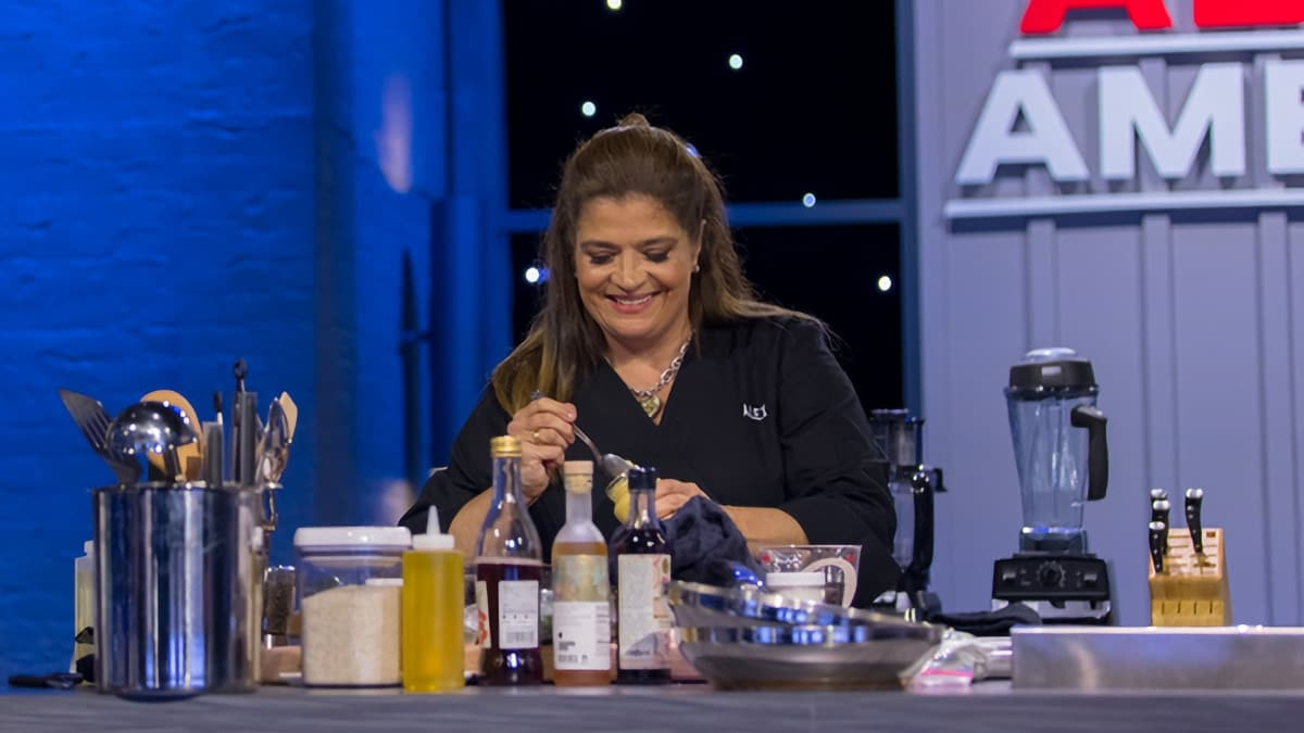 A chef using a spoon to remove something from a jar; the table has several bottles and cooking implements on it